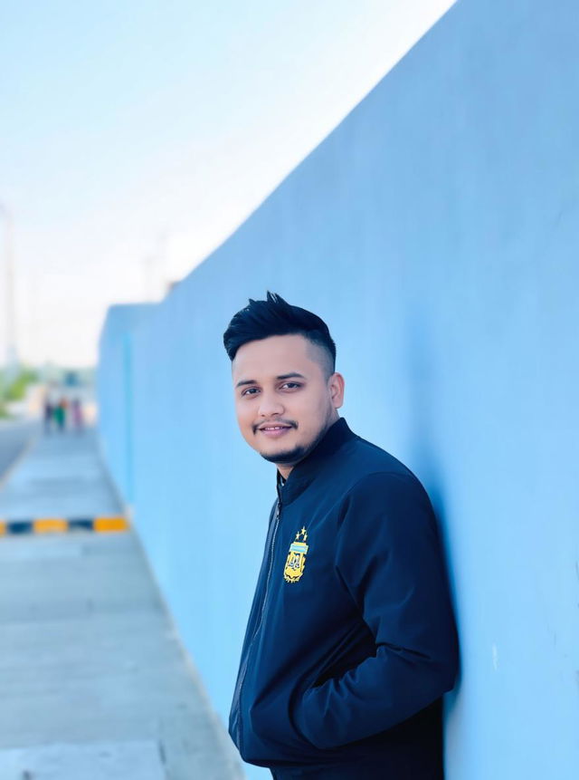 A young man with a charming smile, wearing a stylish black blazer, leaning casually against a bright blue wall in a sunny outdoor setting