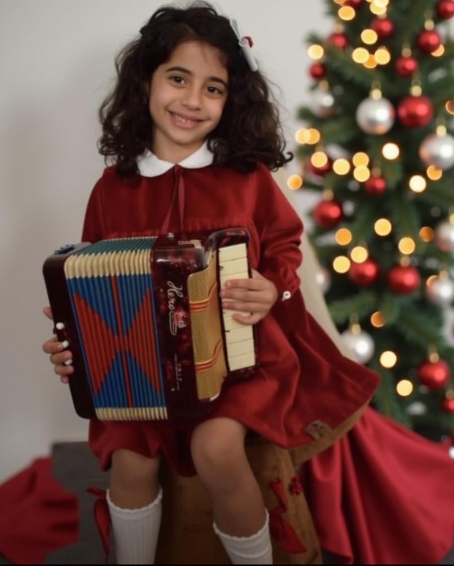 A whimsical Christmas photoshoot scene, featuring a joyful young girl in a vibrant red dress with white accents, sitting on an old wooden chest