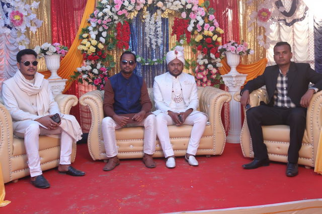 A vibrant Indian wedding scene featuring four well-dressed men seated on elegant sofas adorned with colorful floral decorations