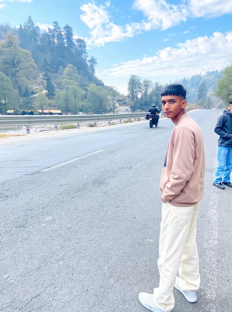 A young man standing on a roadside, wearing a light brown sweater and white pants, with a casual pose