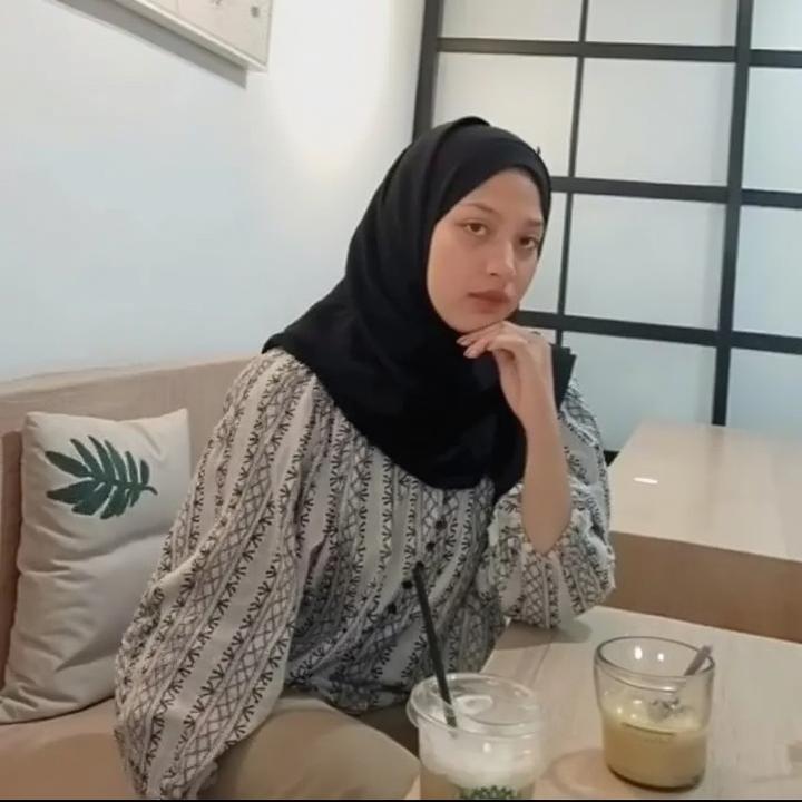 A beautiful woman sitting thoughtfully in a modern cafe, wearing a stylish black hijab and a fashionable, patterned blouse