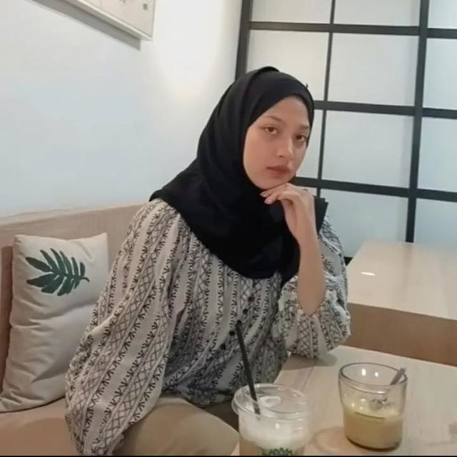 A beautiful woman sitting thoughtfully in a modern cafe, wearing a stylish black hijab and a fashionable, patterned blouse