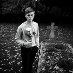 A young man holding a white rose, standing next to an open grave during the autumn season