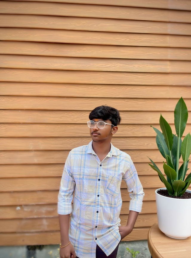 A young man standing confidently against a wooden wall, wearing a checkered shirt and glasses