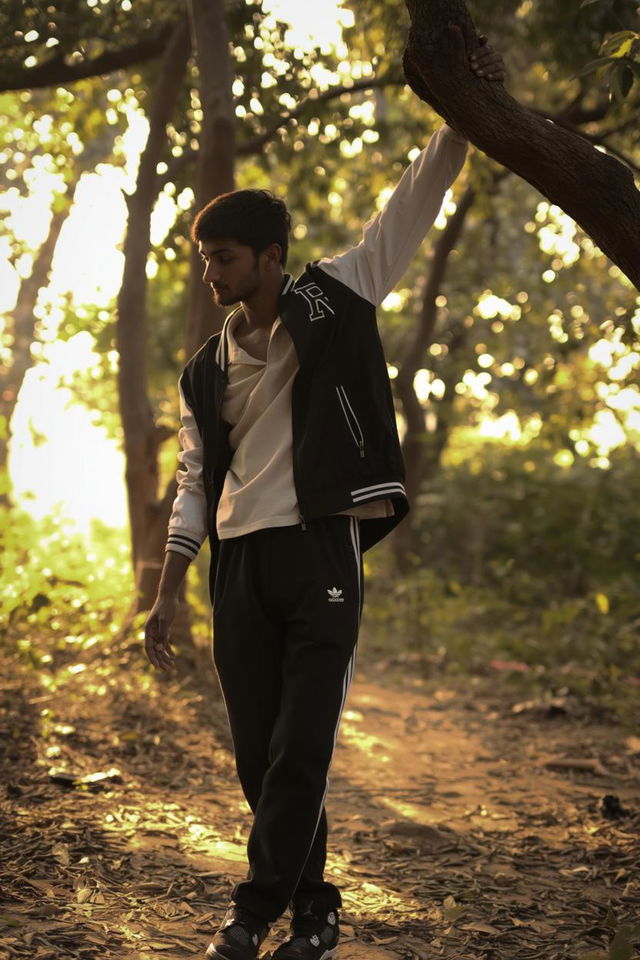 A young man casually dressed in a stylish black varsity jacket and black Adidas track pants, standing relaxed in a lush, green forest setting