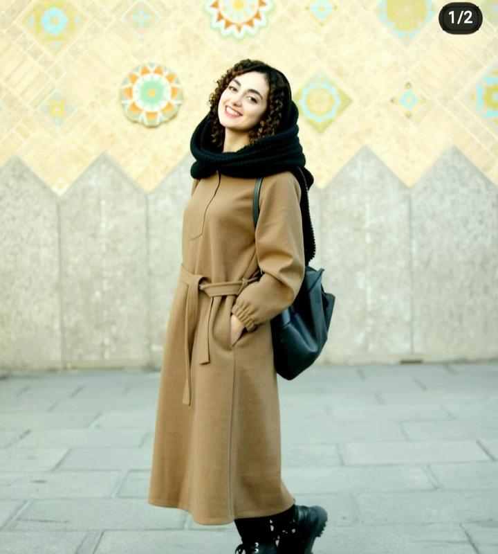 A young woman with curly hair, wearing a stylish, long brown coat with a belt, black scarf, and black boots, standing confidently with a backpack slung over one shoulder