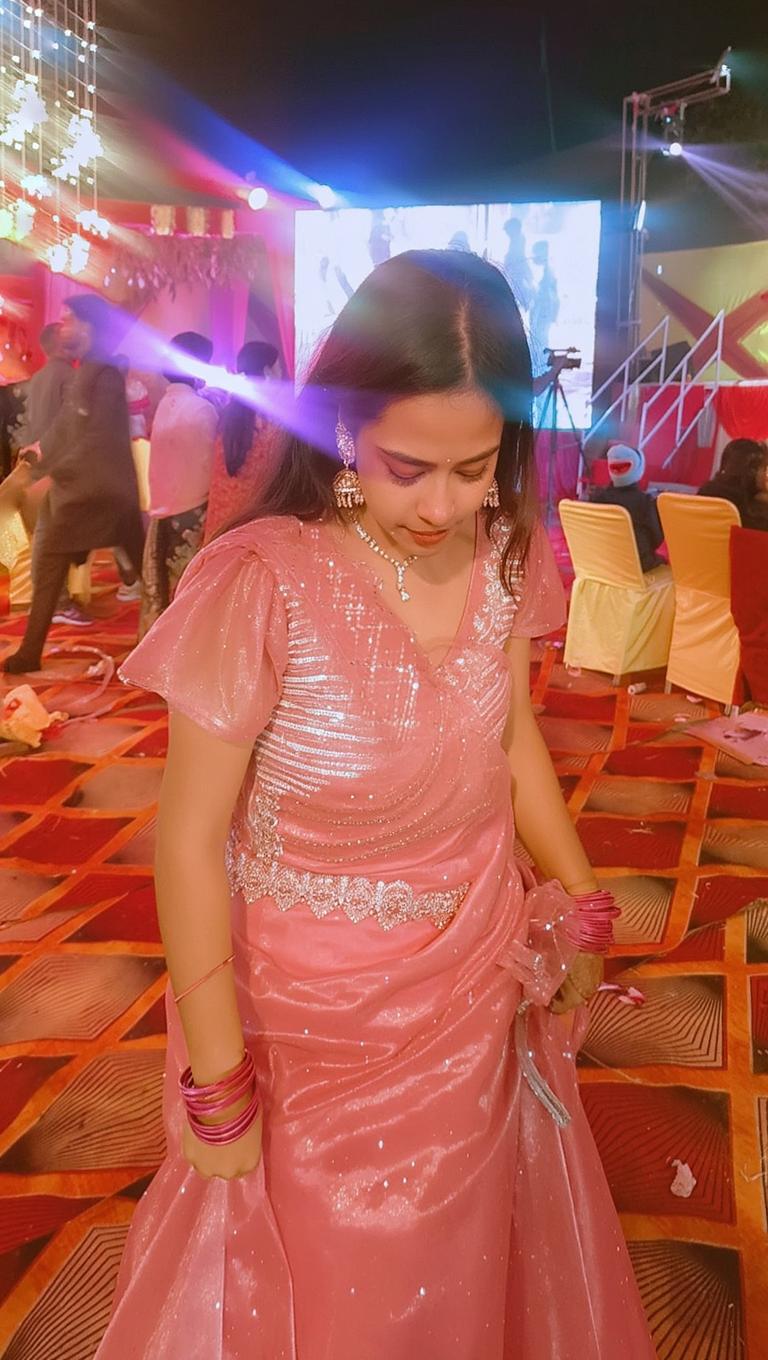 A graceful young woman wearing a beautiful pink traditional outfit with intricate embellishments, captured in a lively party environment