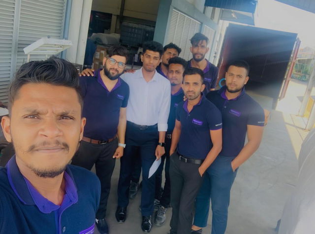 A vibrant group photo of eight professionals in smart navy blue polo shirts with purple accents, standing together in a bright, open warehouse setting