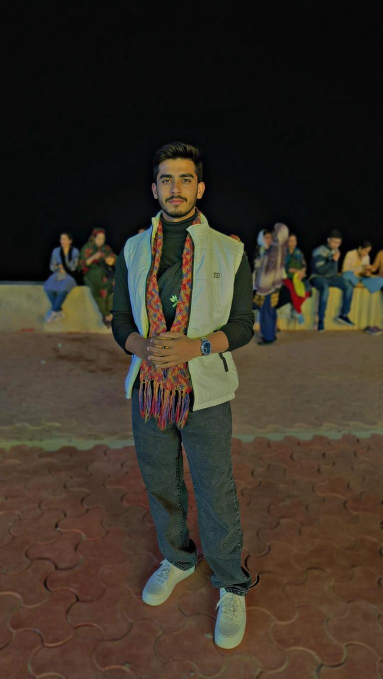 A young man standing confidently in a casual pose, wearing a light-colored vest over a black t-shirt, with a colorful scarf around his neck