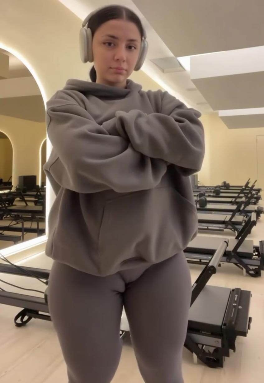 A confident young woman standing in a gym, wearing a large gray hoodie and form-fitting gray leggings