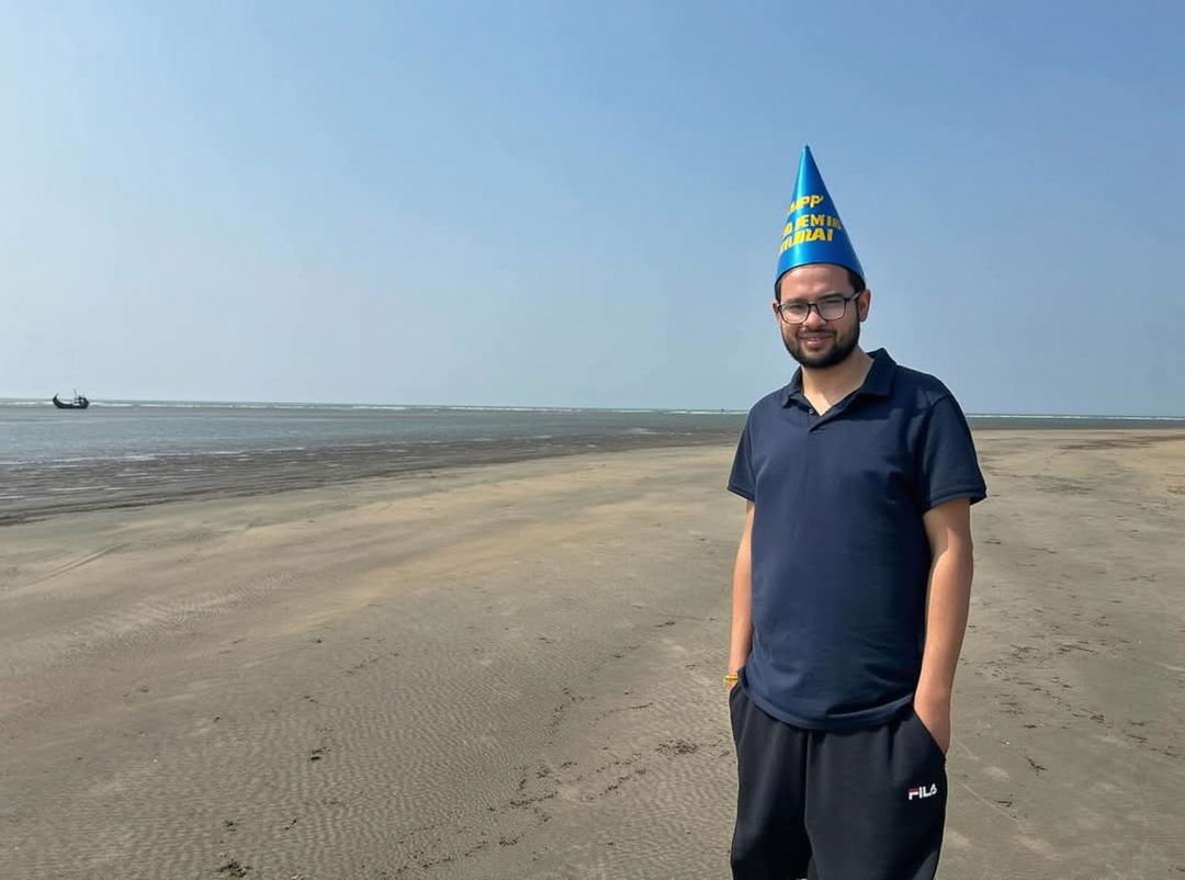 A joyful person standing on a serene beach with a happy birthday hat on their head