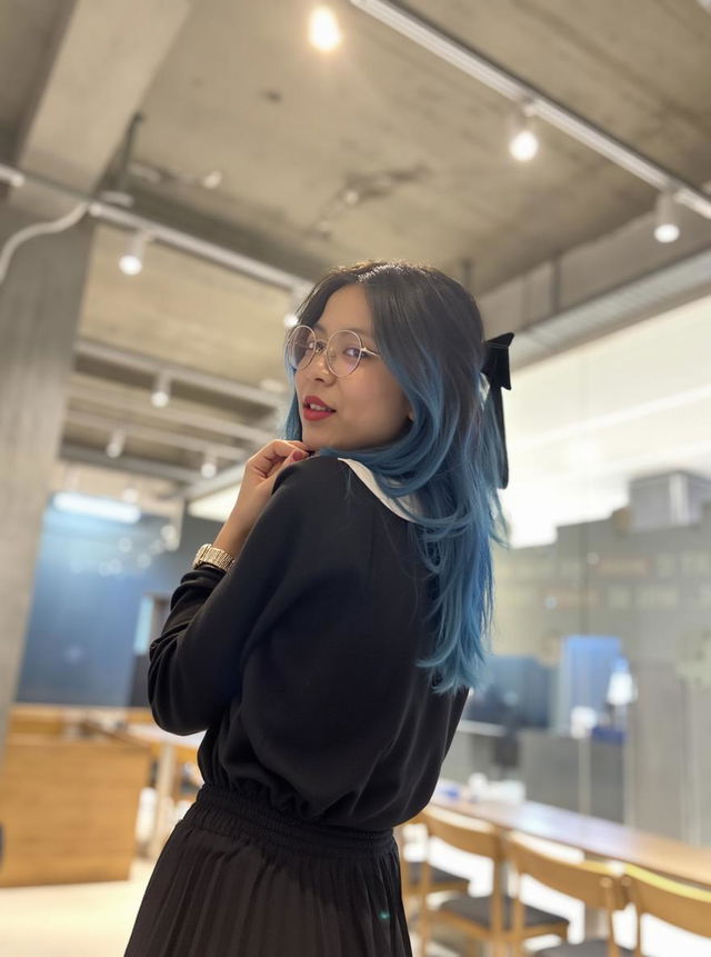 A stylish young woman with long blue hair, wearing a chic black top and a flowing red skirt