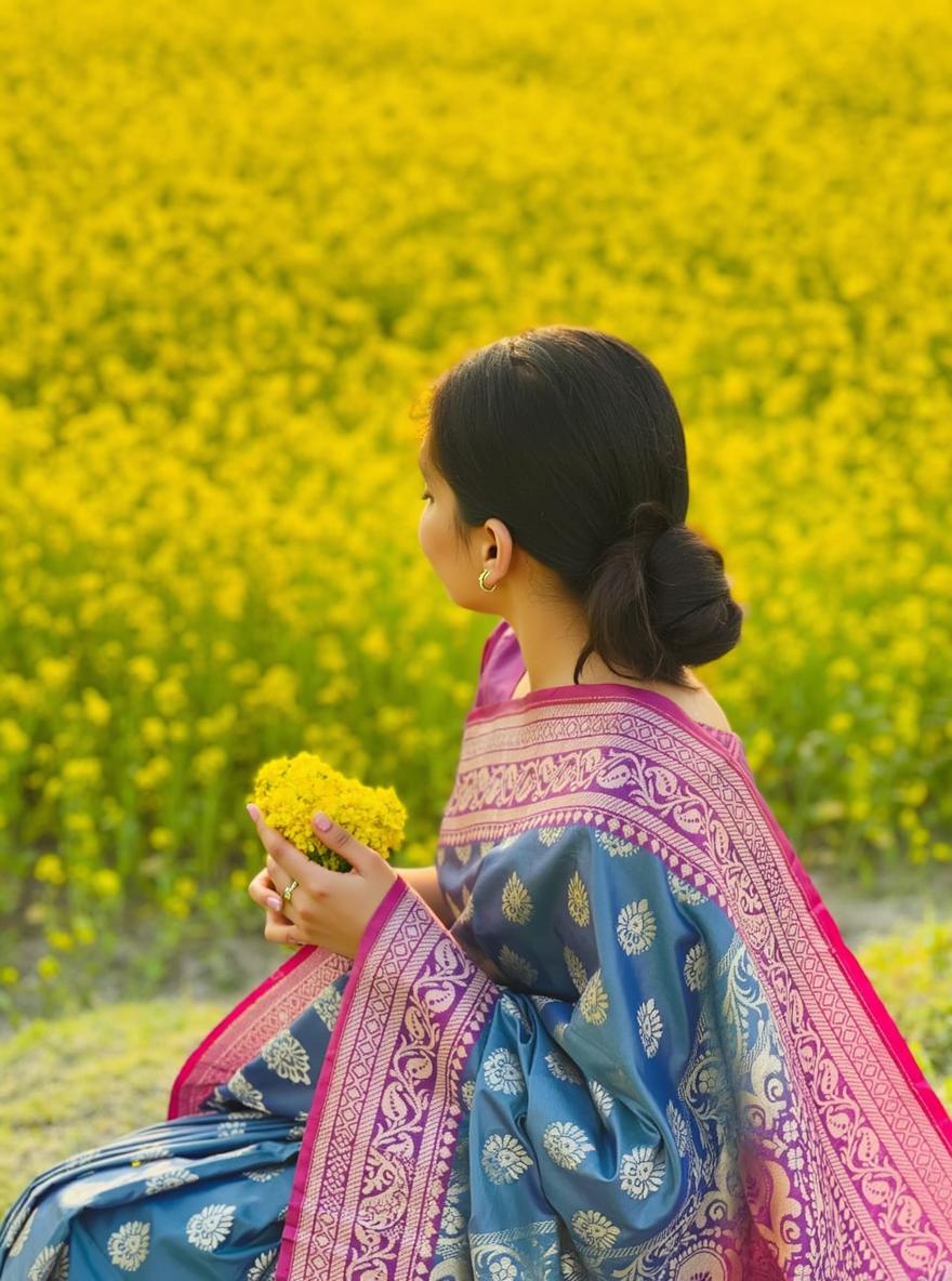 A woman is sitting gracefully in a vast mustard field, wearing a traditional saree that is deep blue with intricate golden motifs, complemented by a stunning pink border