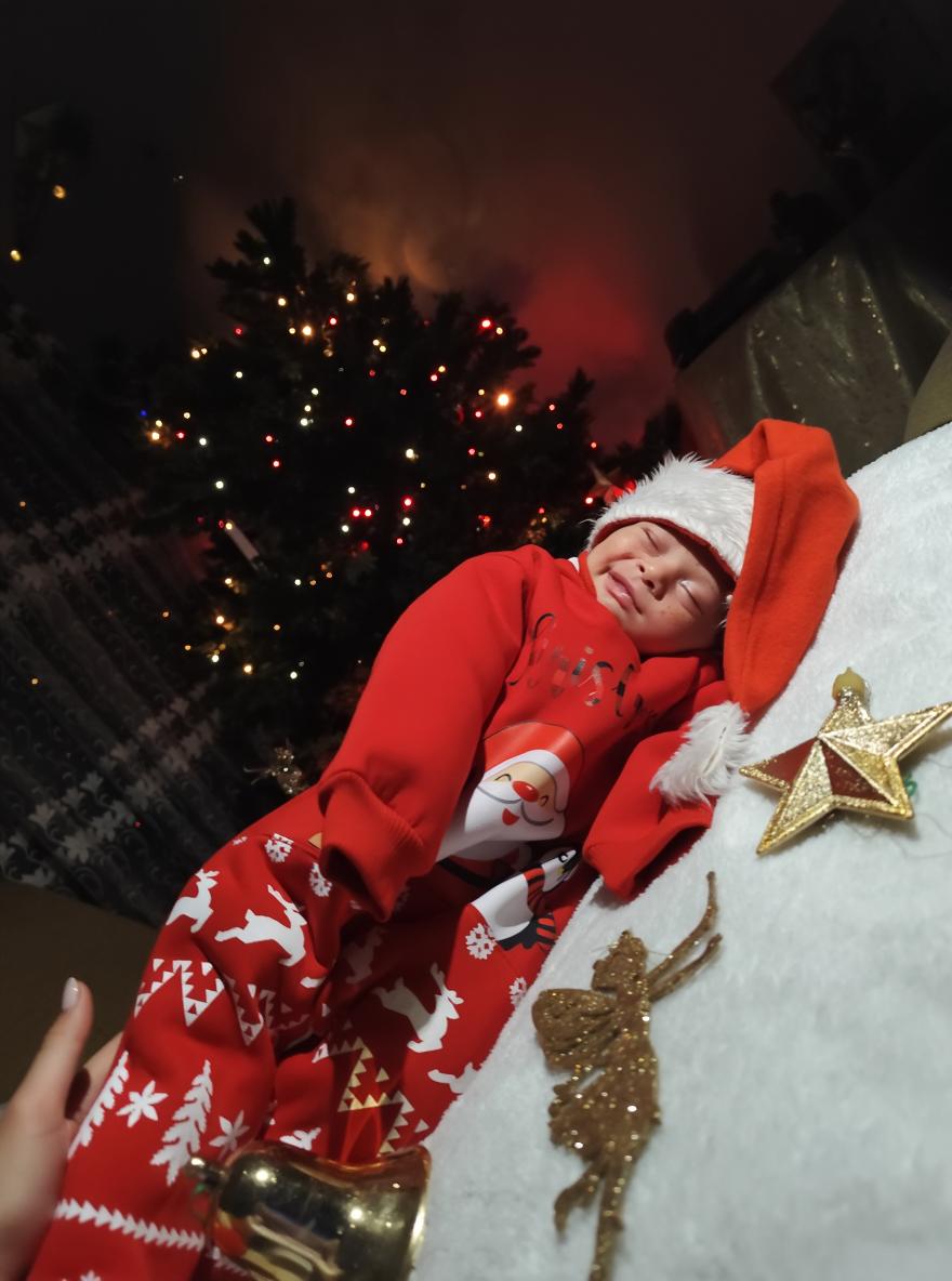 A cozy Christmas scene featuring a smiling baby dressed in a festive red outfit with a Santa hat, lying on a soft white surface adorned with holiday decorations like shiny gold stars, a fairy figurine, and colorful gifts