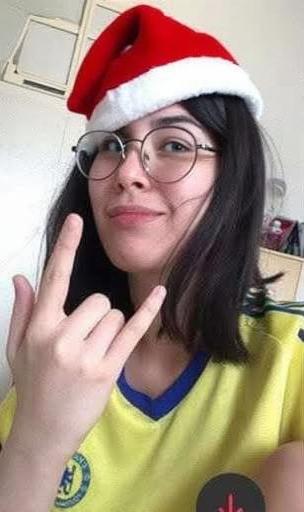 A young woman with shoulder-length black hair and glasses, wearing a yellow sports jersey, making a playful gesture with her hands