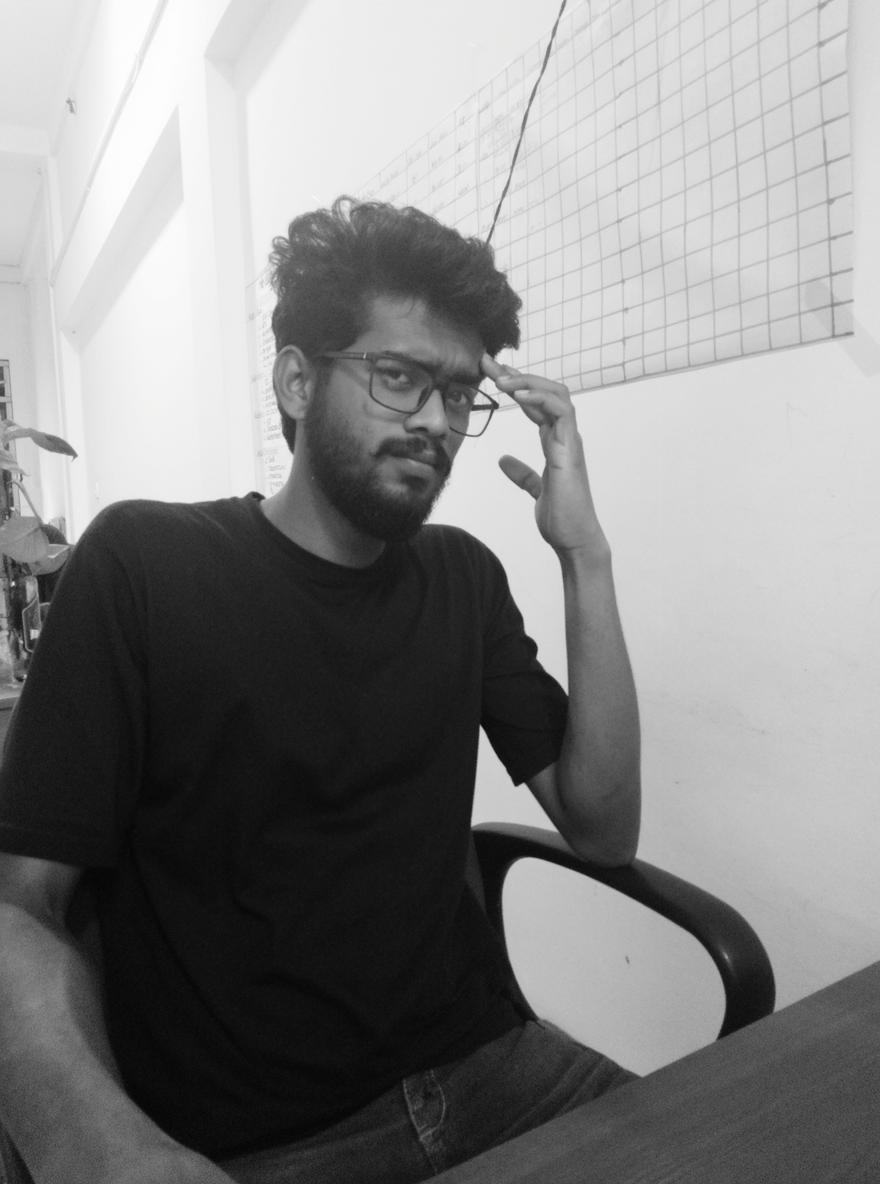 A young man with curly hair and glasses sitting at a desk, looking thoughtfully to the right, hand on his chin