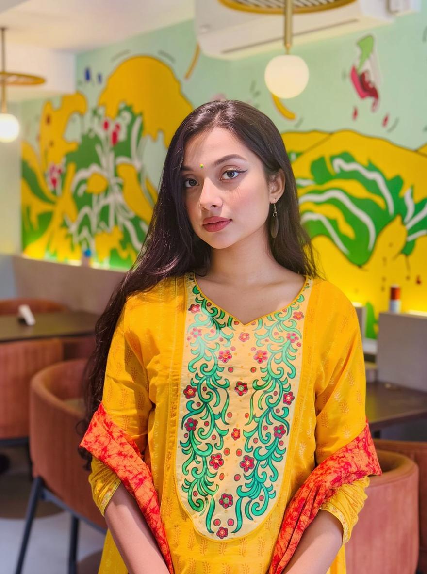 A young woman with long dark hair wearing a vibrant yellow ethnic outfit, intricately designed with colorful patterns and red accents
