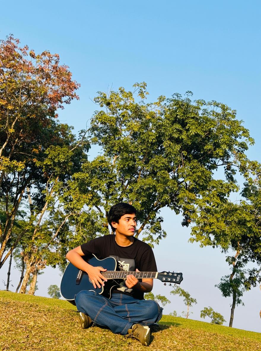 A young man playing an acoustic guitar, sitting cross-legged on a grassy hillside