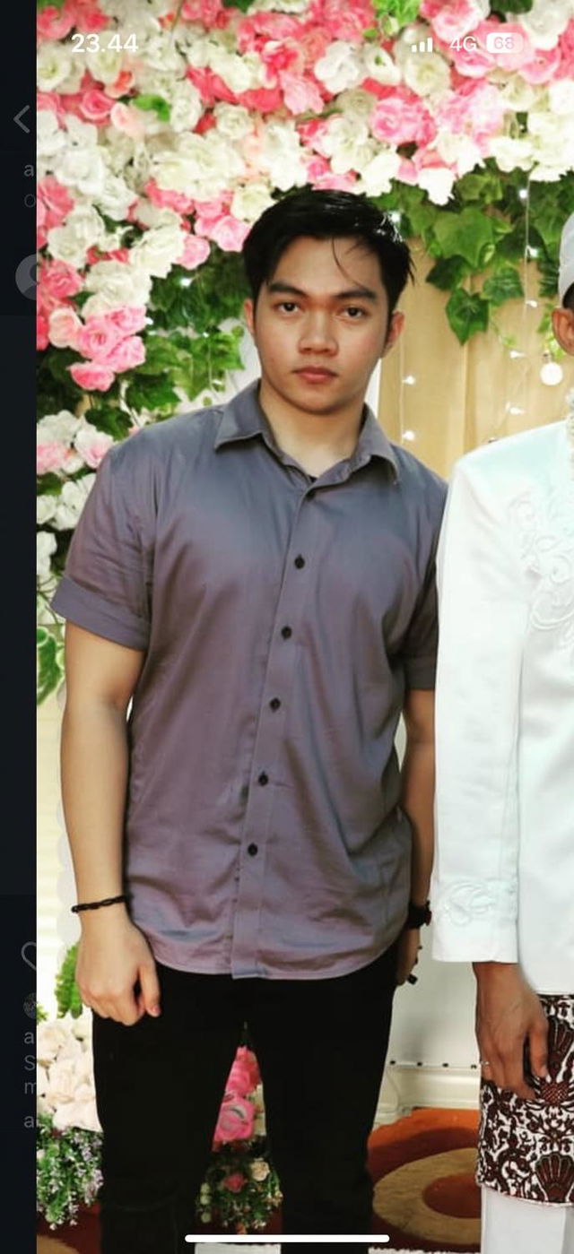 A young man standing, showcasing his left hand prominently without any obstructions, wearing a fitted light gray shirt and black pants