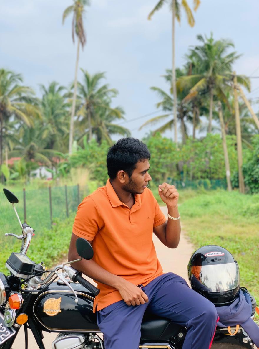 A young man sitting casually on a motorcycle, wearing an orange polo shirt and dark blue pants