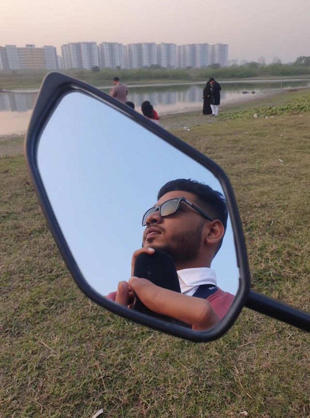 A young man with a stylish beard, wearing sunglasses and a casual outfit, gazes thoughtfully into a mirror