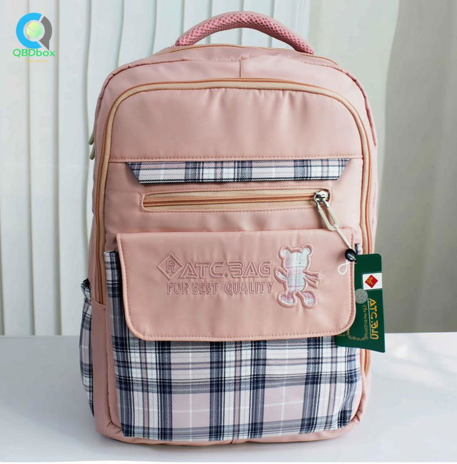 A stylish pink backpack with plaid details at the bottom, featuring a front zip pocket and a teddy bear emblem