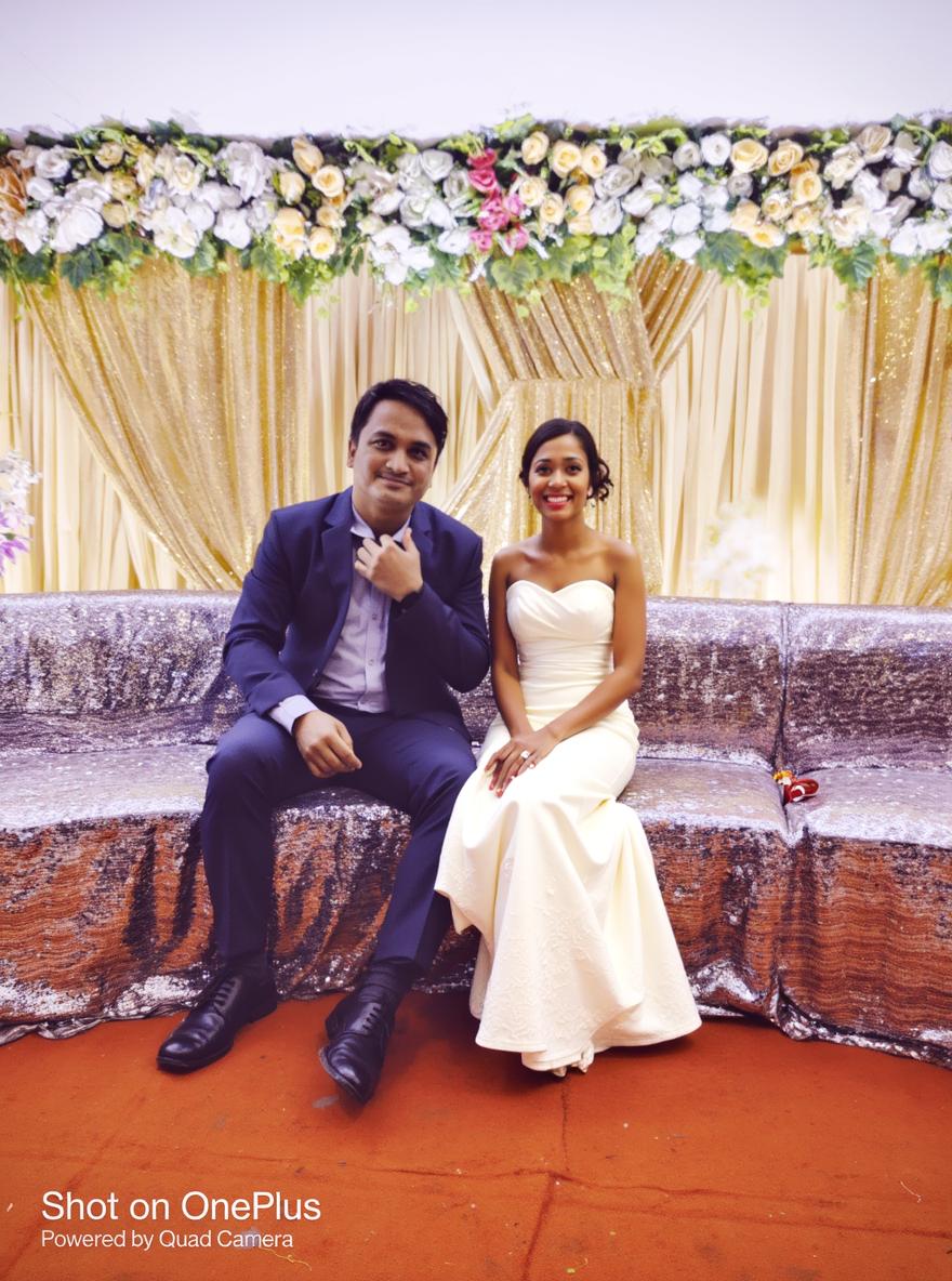 A groom in a formal suit sitting on a decorated sofa with a bride in a traditional wedding outfit, adorned with intricate jewelry and a vibrant bridal lehenga