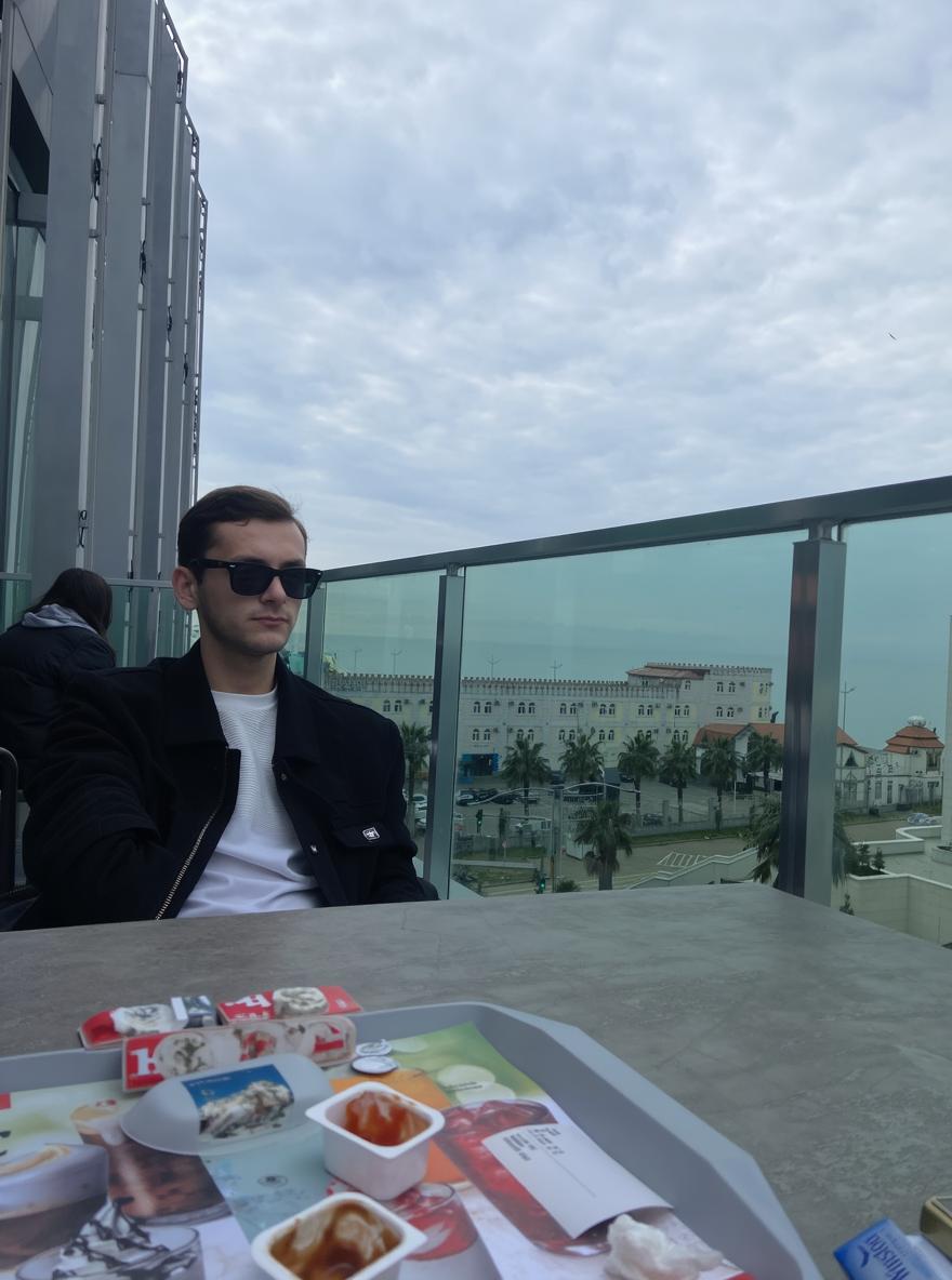 A stylish young man wearing sunglasses, seated at a modern outdoor café on a balcony with a cityscape in the background, featuring cloudy skies and urban architecture