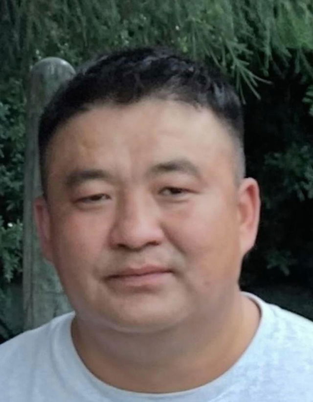 A portrait of a middle-aged Asian man with short dark hair, wearing a casual white T-shirt, standing outdoors with a neutral expression