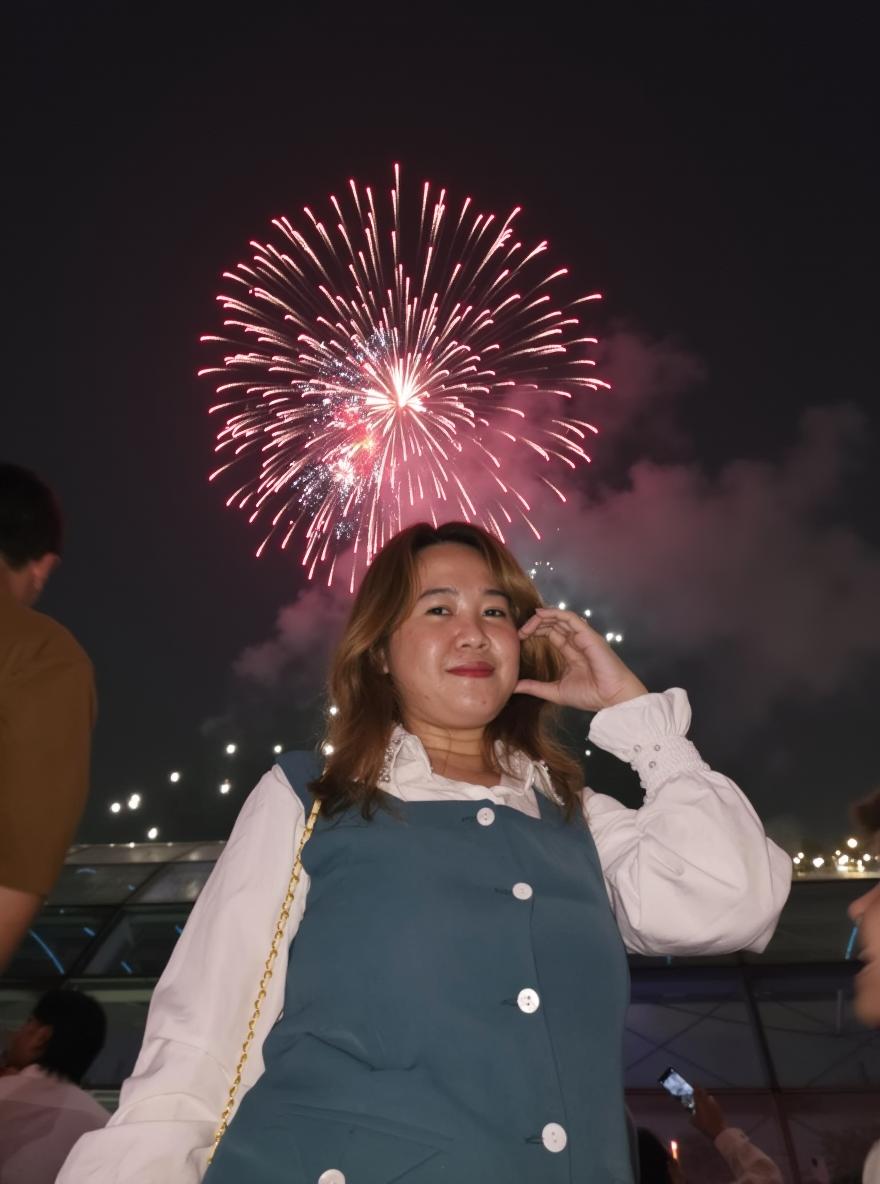 A joyful woman in a stylish teal dress stands against a backdrop of colorful fireworks lighting up the night sky