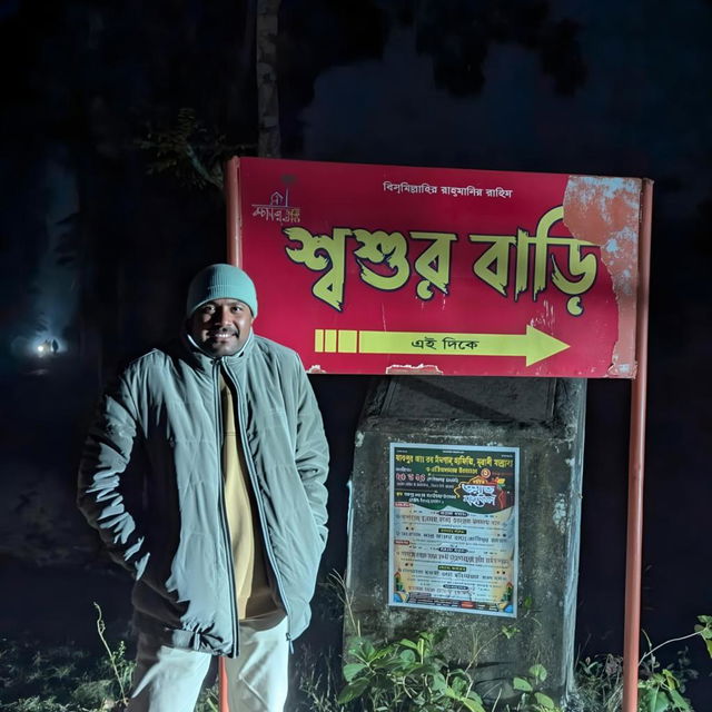 A nighttime scene featuring a young man standing confidently next to a bright red sign which reads 'শুভর বাড়ি', illuminated by soft light