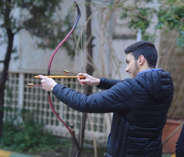 A young man wearing a stylish black puffer jacket is focused on preparing to shoot an arrow from a sleek, traditional crossbow