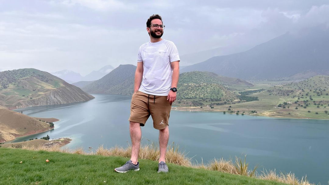 A scenic landscape featuring a man standing confidently on the edge of a grassy hill, overlooking a serene lake surrounded by rolling green hills and mountains