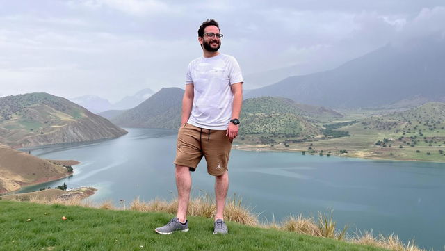 A scenic landscape featuring a man standing confidently on the edge of a grassy hill, overlooking a serene lake surrounded by rolling green hills and mountains