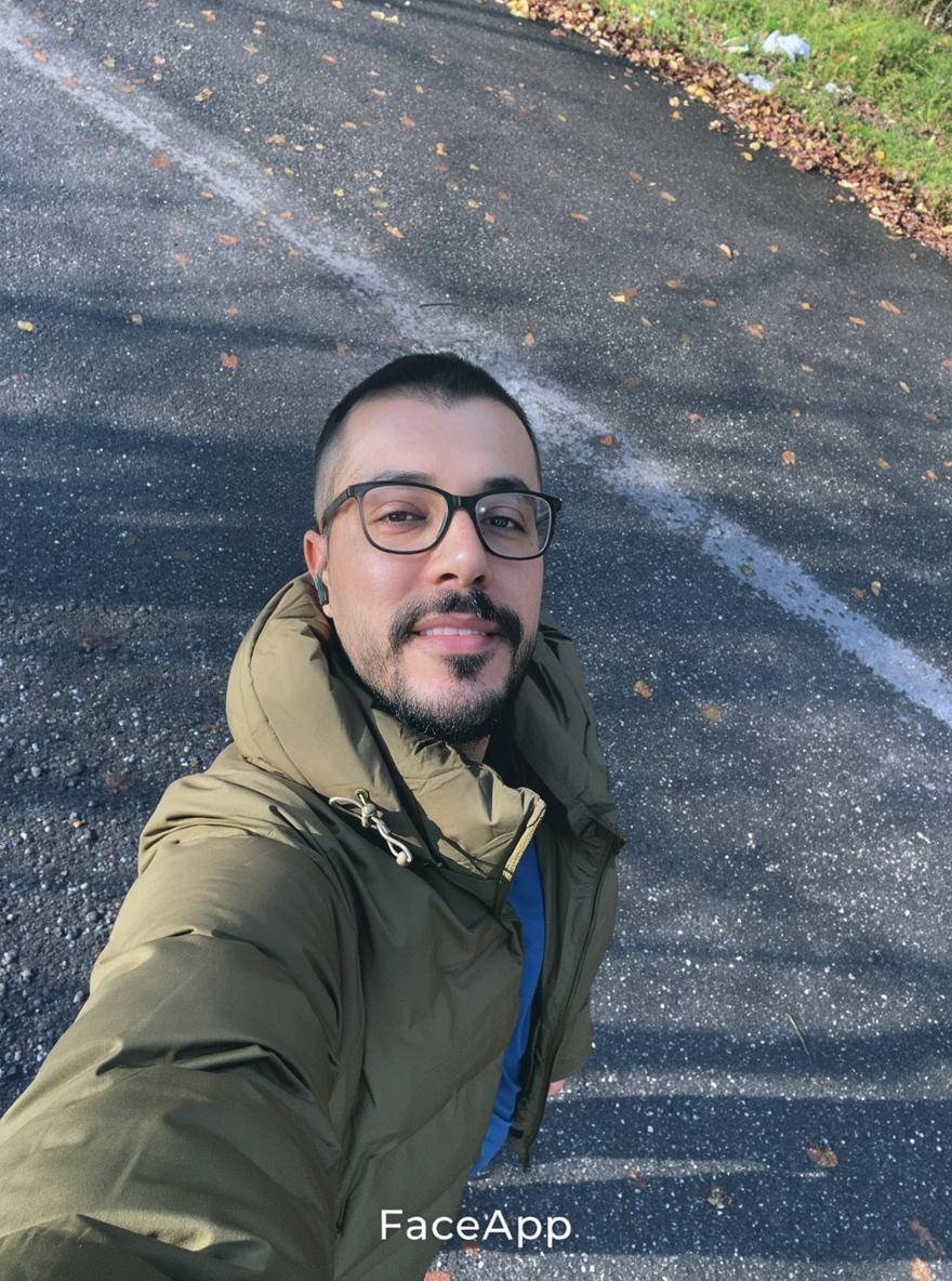 A male figure posing for a selfie in an outdoor setting, wearing a stylish olive green puffer jacket and glasses