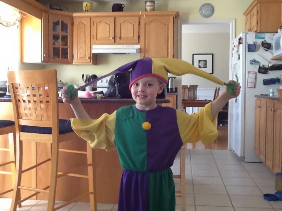 A playful child dressed in a colorful jester costume with a purple and yellow pattern, playfully sticking out their tongue while holding jester props