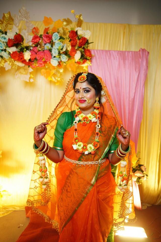 A woman wearing a traditional orange and green saree, adorned with bright floral jewelry and intricate henna designs on her hands
