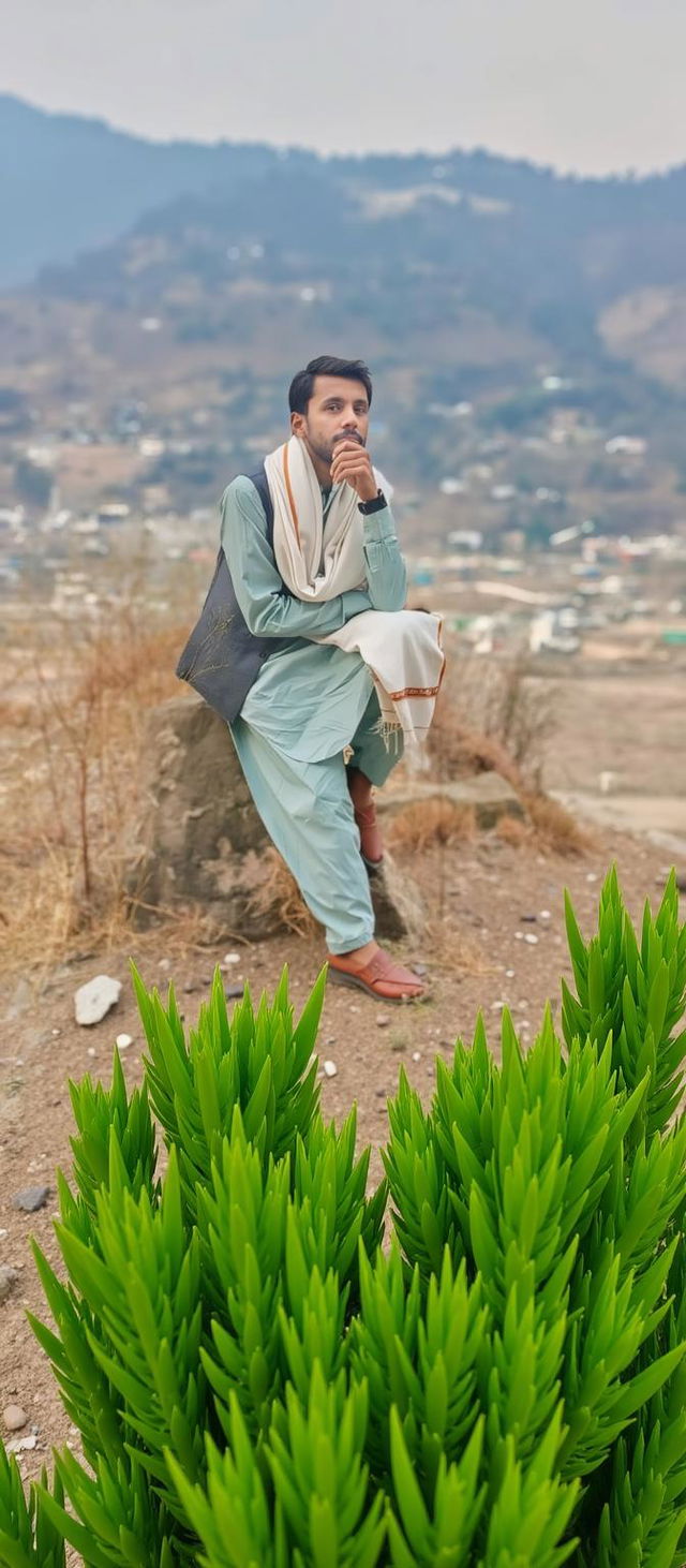 A thoughtful young man dressed in traditional attire, sitting on a rock in a picturesque landscape