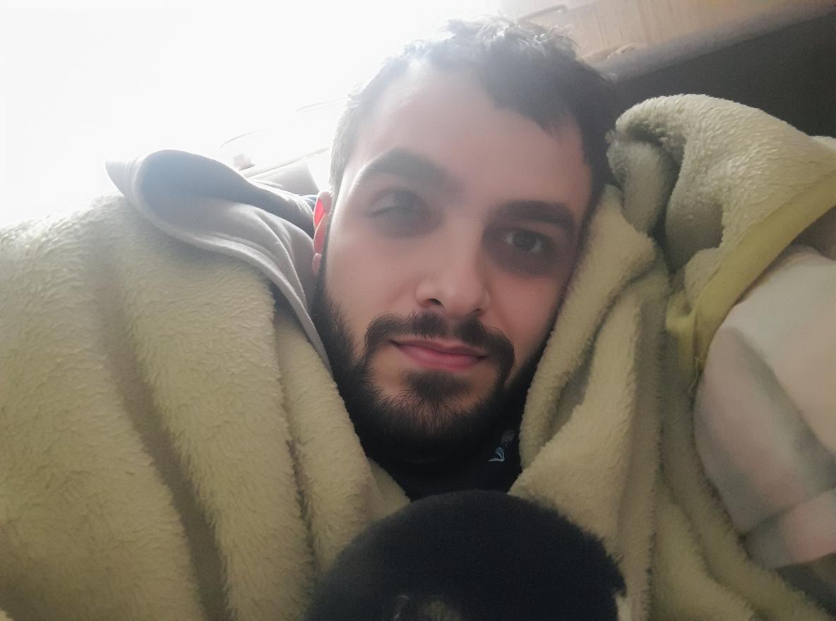 A cozy indoor scene featuring a person wrapped in a soft, fluffy green blanket, with a friendly expression and no beard, cuddling with a small black and white cat