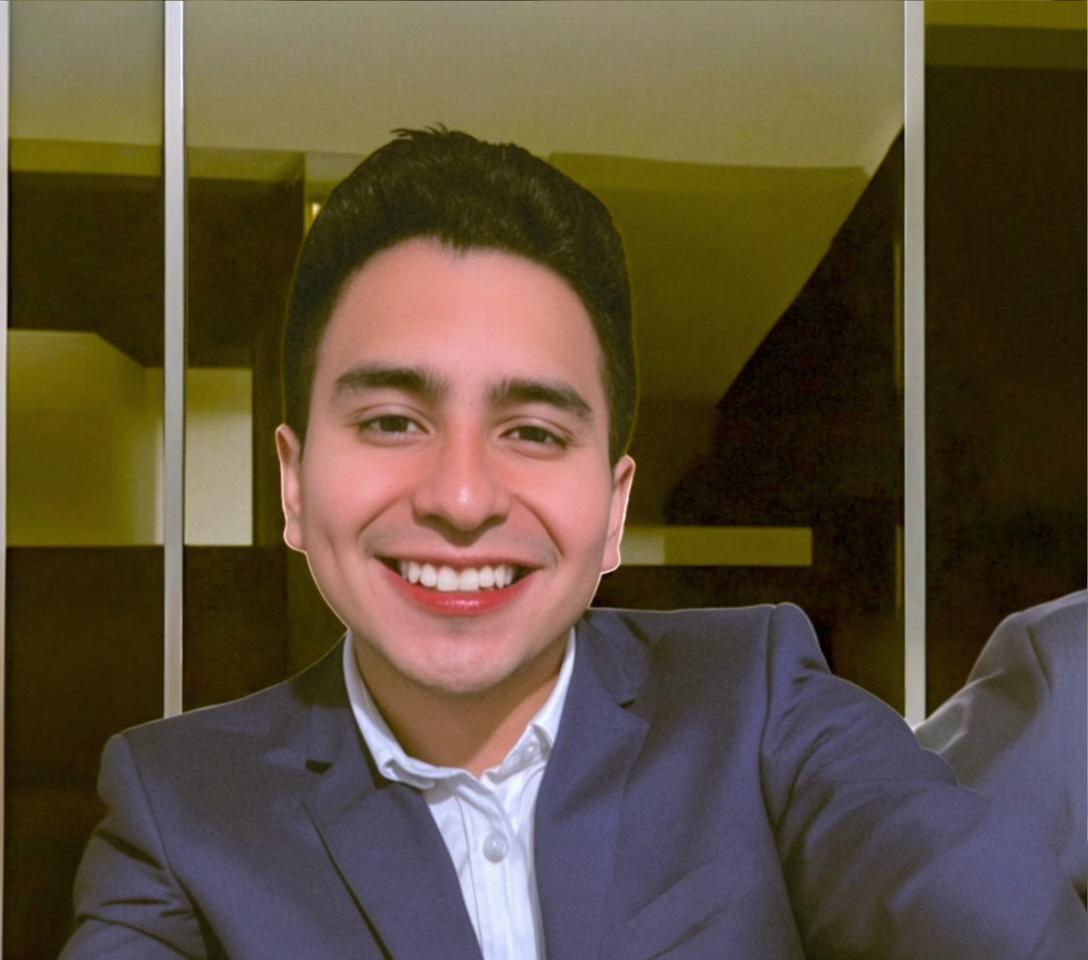 A professional headshot of a young male in a dark suit and white shirt, smiling confidently