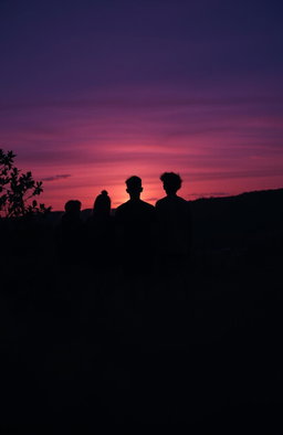 A group of teenagers standing on a hill with their backs turned, silhouetted against a fading sunset