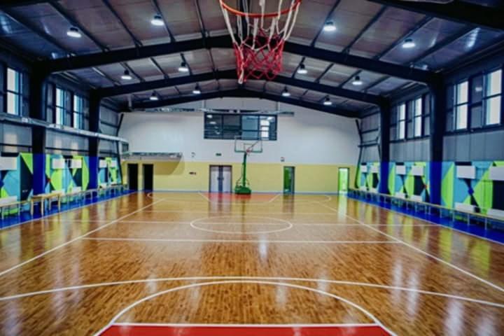 A modern indoor basketball gymnasium featuring a polished wooden floor, vibrant geometric patterns on the walls, large windows allowing natural light to flood in, and a bright, spacious atmosphere