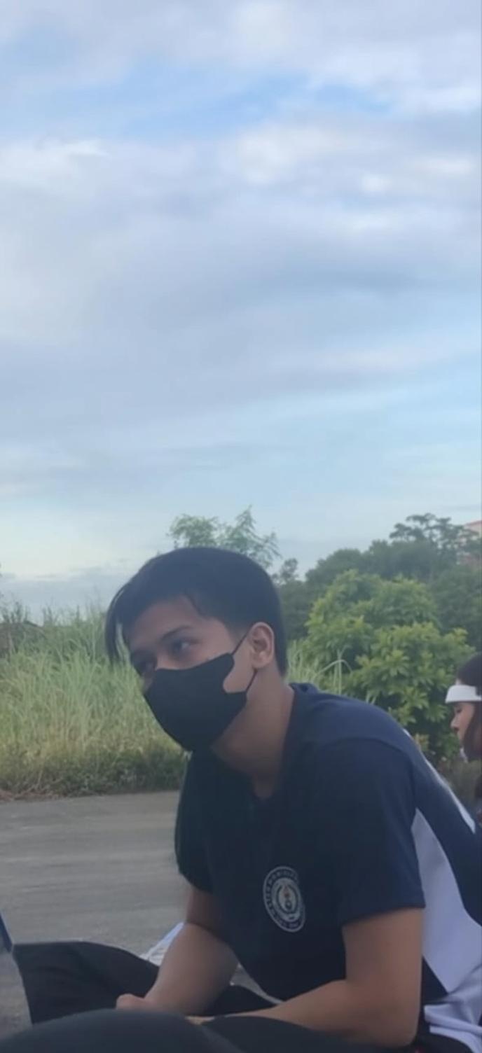 A serene outdoor scene featuring a young individual sitting casually, wearing a black mask and a blue athletic shirt, surrounded by lush greenery and a clear blue sky