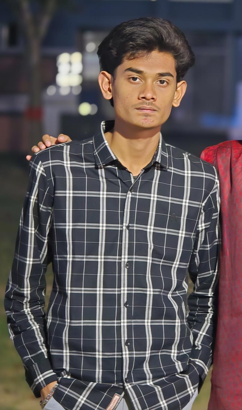 A young man with a serious expression standing confidently in a checkered black and white shirt, hands in his pockets