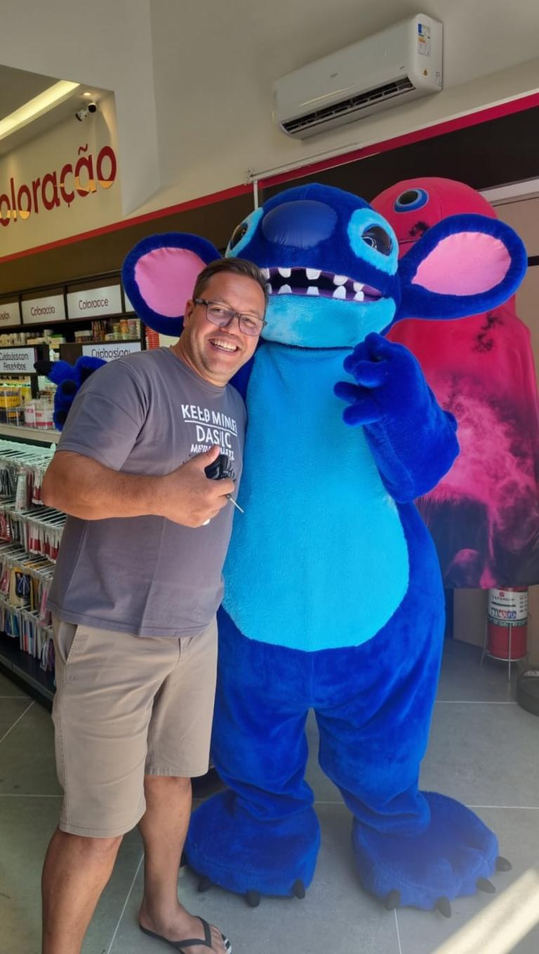 A humorous scene featuring a man with a large, enlarged head, smiling and giving a thumbs up, standing next to a blue stuffed animal character resembling a cartoon alien with a smaller head