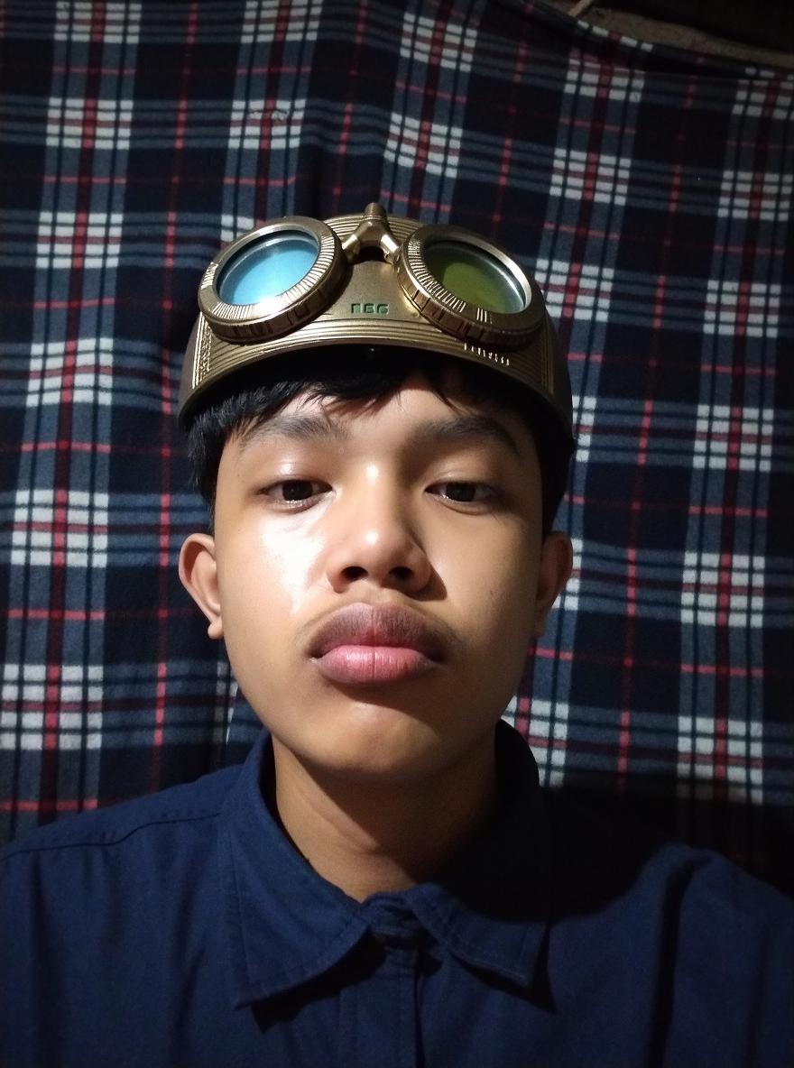 A close-up portrait of a young male wearing an engineering helmet, with a serious expression, set against a background of a plaid patterned fabric