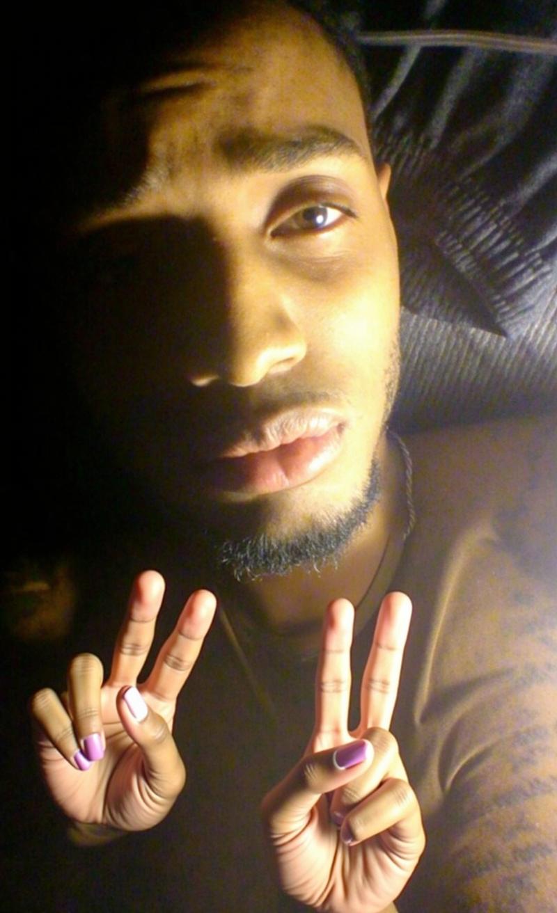 A close-up portrait of a young man lying on a bed, making a peace sign with his right hand in front of his face