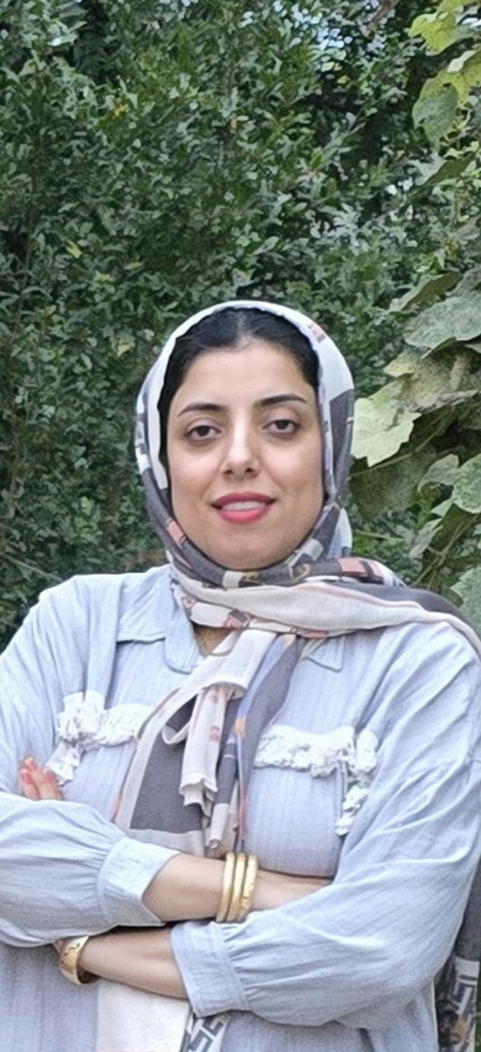 A confident woman wearing a stylish hijab, crossed arms, standing in front of a lush green background filled with leaves and plants