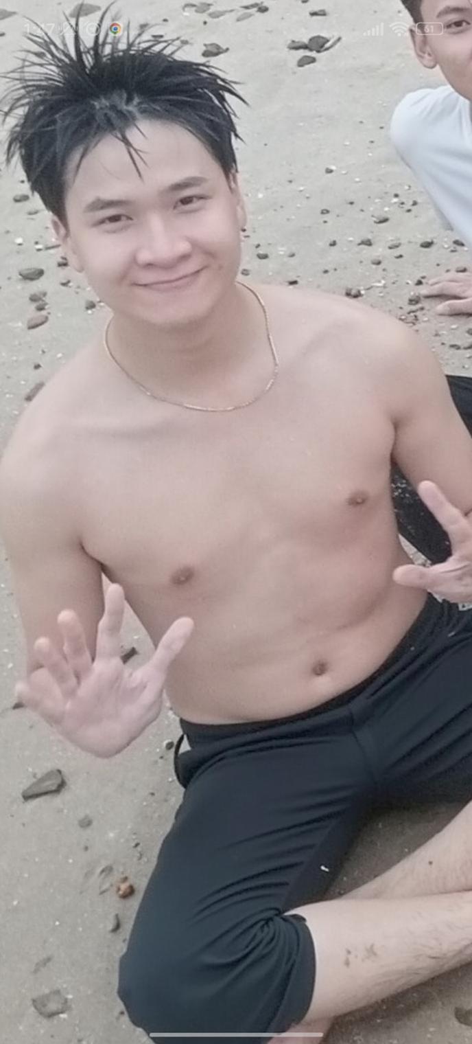 A young man with a charming smile, same face and body as in the image provided, sitting on the beach with a joyful expression
