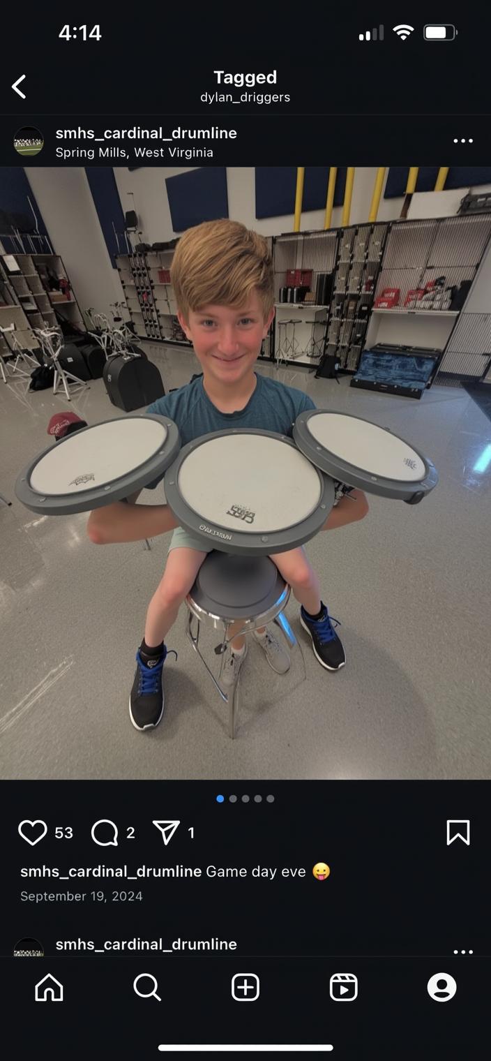 A young boy with short blond hair, smiling and standing in a music room, holding three drum pads